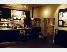 Ein kleiner Verkaufsbereich in einem Kino oder Veranstaltungsort mit einer Theke, die Snacks und Getränke anbietet, beleuchtet durch warme Deckenstrahler. An den Wänden sind Poster angebracht, und eine offene Tür führt in einen weiteren Raum mit Backsteinwand.