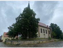 Eine kleine historische Kirche mit hellen Wänden, dunklen Fenstern und einem spitzen Kirchturm, umgeben von großen Bäumen. Der Platz vor der Kirche ist mit Kopfsteinpflaster belegt, und eine niedrige Steinmauer umgibt das leicht erhöhte Gelände.