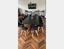 Ein moderner Besprechungsraum mit einem schwarzen Tisch, umgeben von schwarzen Stühlen mit Holzbeinen, auf einem Fischgräten-Holzboden. Im Hintergrund stehen Regale mit Geschirr, Getränken und Geräten, sowie ein großer Flachbildfernseher an der weißen Wand.