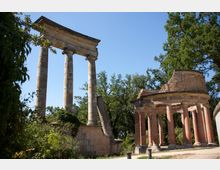 Ruinen eines historischen Tempels mit dorischen Säulen und einem runden, teilweise zerstörten Säulengebäude aus Ziegelstein, umgeben von Bäumen und Vegetation bei sonnigem Himmel.