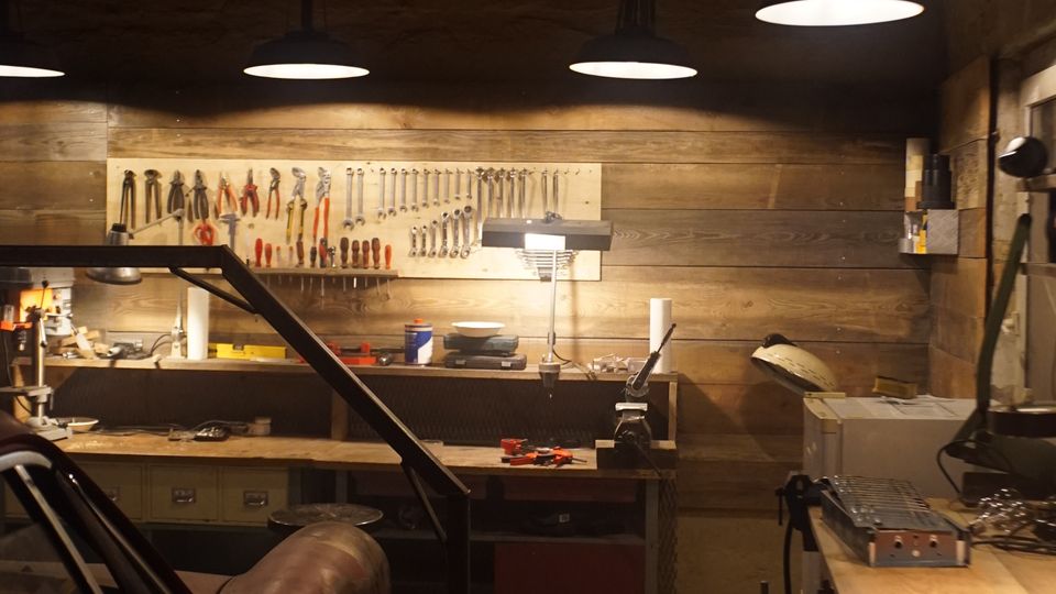 Innenansicht einer Werkstatt mit Holzvertäfelung, großer Werkbank und einer Werkzeugwand mit aufgehängten Schraubenschlüsseln und Zangen. Mehrere Pendelleuchten hängen von der dunklen Decke, daneben stehen Werkstattgeräte und links ist ein teilweise sichtbares Fahrzeug oder Maschinengehäuse.