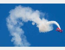 Ein rotes Kunstflugzeug erzeugt eine weiße Rauchspur, während es in einer Kurve am strahlend blauen Himmel fliegt.