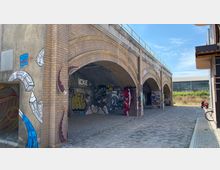 Architektonische Bogengänge aus Backstein mit Graffiti an den Wänden unter einer erhöhten Struktur. Der Bereich ist mit Pflastersteinen bedeckt, und im Hintergrund befinden sich ein modernes Gebäude sowie eine Grünfläche.