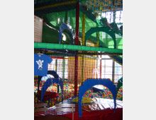 Indoor-Spielplatz mit bunten Spielgeräten, Netzen und einer großen Ballgrube. Im Vordergrund sind Sitzbänke und ein blauer Stoff mit einem Motiv aufgehängt.