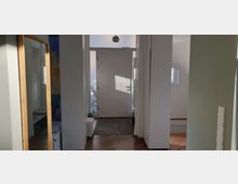 Ansicht eines modernen, hellen Eingangsbereichs eines Hauses. Zu sehen sind eine weiße Haustür mit Glaselementen, ein Holzboden in Fischgrätenmuster, ein großer Wandspiegel links und weiße Einbauschränke auf beiden Seiten.