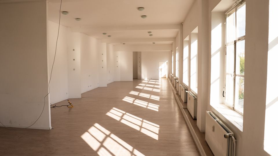 Langer, leerer Innenraum mit weißen Wänden, hellem Holzboden und einer Reihe großer Fenster entlang der rechten Seite. Sonnenlicht fällt durch die Fenster und wirft rechteckige Schatten; unter den Fenstern stehen Heizkörper.