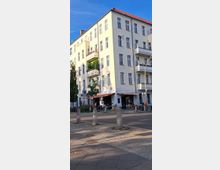 Ecke eines mehrstöckigen Wohngebäudes mit hellgelber Fassade, Balkonreihen und rotem Dach. Im Erdgeschoss befindet sich ein Café mit Außenbestuhlung, umgeben von Bäumen, Fahrrädern und einer gepflasterten Gehwegfläche entlang einer ruhigen Straße.