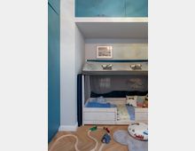 Kinderzimmer mit eingebautem Hochbett aus Holz, dekoriert mit Dinosauriermotiv-Vorhängen. Im Vordergrund liegt eine Holzeisenbahn auf einem hellen Parkettboden, umgeben von einem runden Spielkissen und einem grauen Teppich.