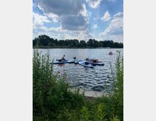 Ein See mit mehreren Menschen auf Stand-Up-Paddle-Boards; im Vordergrund sind Gräser und Blumen zu sehen, während im Hintergrund Bäume und ein bewölkter Himmel erkennbar sind.