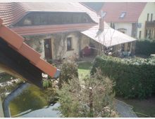 Wohnhof mit einem gelben Gebäude und rotem Ziegeldach im ländlichen Stil. Im Vordergrund ein Teich mit Fischen, umgeben von Pflanzen, sowie eine grüne Hecke und ein gepflasterter Pfad.