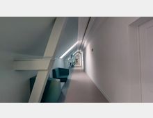 Langer, schmaler Flur mit schrägen Dachbalken und modernem Design. Auf der linken Seite befinden sich Sitzbänke, die in die Nischen eingefügt sind, und die rechte Wand ist glatt und weiß gestrichen, mit einer Notausgangsbeschilderung.