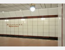 U-Bahn-Station mit weiß gefliester Wand, auf der in roten Buchstaben "Altstadt Spandau" steht. Der Boden ist mit rötlich-beigen Fliesen ausgelegt, und eine runde Deckenleuchte hängt zentral über dem Bereich.