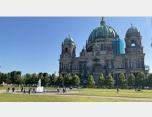 Eine historische Kathedrale mit großen grünen Kuppeln und verziertem Fassadenschmuck, umgeben von Bäumen und einer Wiese. Im Vordergrund befindet sich ein Brunnen, bei dem Menschen spazieren und die Umgebung genießen.