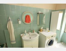 Ein Badezimmer mit mintgrünen Fliesen an den Wänden, einem runden Wandspiegel über einem weißen Waschbecken und einem Waschbeckenunterschrank. Rechts steht eine Waschmaschine, daneben ein Regal mit verschiedenen Hygieneartikeln, und an der Wand hängt ein Handtuch.