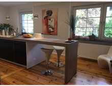 Moderne Küchenansicht mit einer Kücheninsel, zwei weißen Barhockern und einem hölzernen Boden. Große Fenster mit Blick ins Grüne, Pflanzen auf der Fensterbank und ein abstraktes Gemälde an der Wand runden den Raum ab.