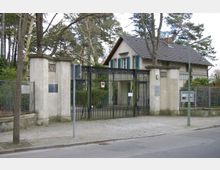 Eingangstor mit hohem Metallzaun und Steinpfosten vor einem Haus mit Satteldach und grünen Fensterläden, umgeben von Bäumen und einem Gehweg. Schilder und Gedenktafeln sind an den Steinpfosten angebracht.