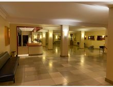 Ein heller Eingangsbereich oder Lobby mit gefliesten Böden, beigefarbenen Wänden und Säulen. Im Vordergrund befindet sich eine schwarze Sitzbank, links ein Empfangstresen mit rotem Rahmen, und an den Wänden hängen dekorative Kunstwerke.