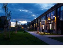 Industriegebäude mit großen Fenstern und Backsteinfassade, beleuchtet bei Dämmerung. Im Vordergrund ein Gehweg, gesäumt von neu gepflanzten Bäumen und Rasenflächen.