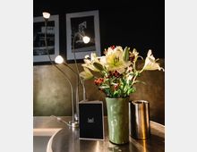 Ein minimalistischer Innenraum mit einem Metalltisch, darauf ein grüner Blumenstrauß in einer Vase, ein silberner Behälter und eine klappbare Karte mit der Aufschrift „Lust.“ Im Hintergrund eine goldfarbene Wand und zwei Schwarz-Weiß-Fotografien.