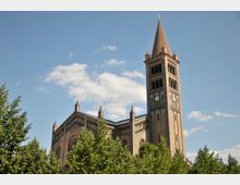 Backstein-Kirche mit einem hohen Glockenturm und spitzem Dach, umgeben von grünen Bäumen, vor einem blauen Himmel mit einigen weißen Wolken.