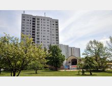 Ein modernes Hochhaus mit mehreren Etagen steht inmitten einer grünen Parklandschaft mit Bäumen und Rasen. Im Vordergrund ist ein kleines Gebäude mit geschwungener Dachform zu sehen, das von einer Werbetafel mit der Aufschrift "Sandbar" geschmückt ist.