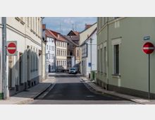 Schmale Stadtstraße mit zweigeschossigen Wohnhäusern aus heller Putzfassade auf beiden Seiten. Verkehrsschilder "Einfahrt verboten" an beiden Straßenseiten, geparkte Autos und vereinzelte Personen im Hintergrund.