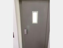 Graue Tür mit rechteckigem Fenster in der oberen Hälfte, das von einem weißen Rahmen umgeben ist. Links an der Tür befindet sich ein senkrechter Metallgriff.