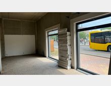 Ein leerer Innenraum mit Betonwänden und einem Boden aus Holzfliesen. Zwei große Fensterfronten mit Blick auf die Straße, wo ein gelber Stadtbus sowie Fußgänger zu sehen sind; an einer Säule im Raum sind gestapelte Dämmmaterialien angelehnt.