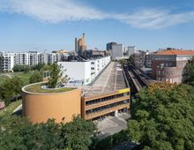 Luftaufnahme eines urbanen Viertels mit modernen Wohngebäuden, einem mehrstöckigen Parkhaus mit begrüntem Dach und einer erhöhten U-Bahn-Trasse. Im Hintergrund sind weitere Büro- und Wohngebäude zu sehen, umgeben von Bäumen und Grünflächen.