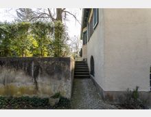 Seitenansicht eines beige verputzten Gebäudes mit grünen Fensterläden und einem gewölbten Kellerfenster. Im Vordergrund befindet sich eine alte Betonmauer mit moosigen Spuren, dahinter ein kleiner Treppenaufgang, der zu einem Gartenbereich mit Bäumen und Hecken führt.