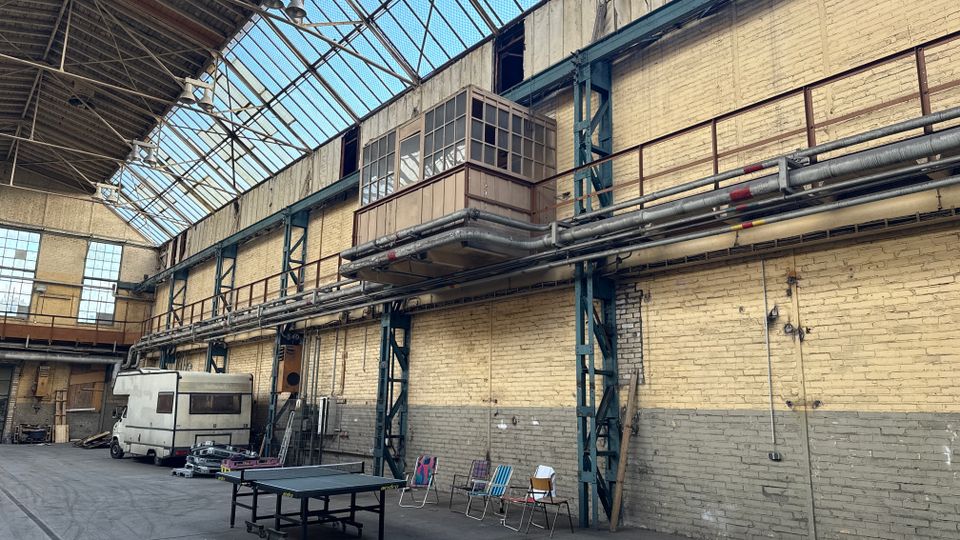 Große Industriehalle mit hohen Backsteinwänden, Stahlträgern und einem durchgehenden Glasdach, das viel Tageslicht hereinlässt. Entlang der Wand verlaufen Rohrleitungen und eine erhöhte Laufbahn; am Boden stehen ein Wohnmobil, ein Tischtennistisch und einige Stühle.