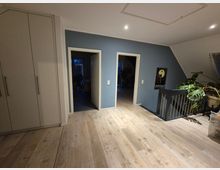 Flur in einem modernen Wohnhaus mit Holzfußboden, blauer Wandfarbe und drei Türen, die zu angrenzenden Räumen führen. Links befindet sich ein großer, weißer Einbauschrank, und rechts ist eine Treppe mit schwarzem Geländer sowie eine Zimmerpflanze sichtbar.