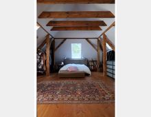 Dachgeschoss-Schlafzimmer mit sichtbaren Holzbalken, einem niedrigen Bett in der Mitte, flankiert von Nachttischen und Lampen. Ein großer, bunt gemusterter Teppich liegt auf dem Holzboden, und an den Seiten befinden sich offene Kleiderablagen sowie ein kleines Regal.