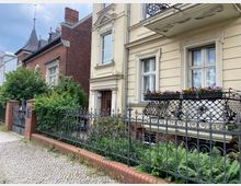 Traditionelle Stadthäuser mit verzierten Fassaden, eines aus rotem Backstein mit Türmchen und eines in hellbeigem Putz mit Fenstern, schmiedeeisernem Balkon und Blumenkästen. Im Vordergrund ein gepflasterter Gehweg, ein Zierzaun mit Backsteinsockel und üppige Pflanzen.