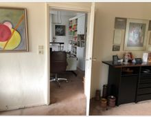 Ein Blick in ein Büro oder Arbeitszimmer, gesehen durch eine offene Tür. Im Vordergrund befinden sich eine schwarze Kommode, mehrere Wandbilder, und ein abstraktes Gemälde an der linken Wand. Im Hintergrund steht ein Schreibtisch mit einem Drehstuhl sowie Regale voller Bücher an der Wand.