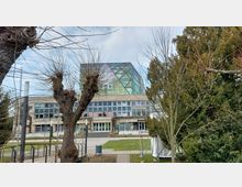 Modernes Gebäude mit einer gläsernen, pyramidenförmigen Dachkonstruktion, unterhalb einer Fassade aus mehreren Fensterreihen. Im Vordergrund stehen kahle und grüne Bäume sowie ein kleiner gepflasterter Weg.