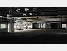 Ein leerer, großzügiger Innenraum einer Industriehalle mit sichtbaren Deckeninstallationen, mehreren Fenstern entlang der Seitenwände und offenen Türen, die in weitere Räume oder Bereiche führen. Die Beleuchtung ist gedämpft, und die Fläche wirkt verlassen.