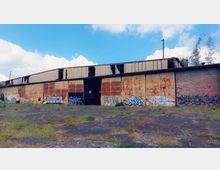 Eine verfallene Industriehalle mit rostigen Metallwänden und einer Backsteinfassade. Die Fassade ist mit zahlreichen Graffitis bemalt, und der Boden davor ist von Gras und Unkraut durchzogen.
