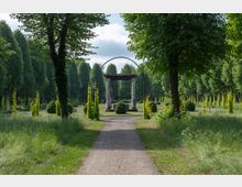 Grüne Parklandschaft mit einem Kiesweg, der auf eine Pergola mit vier weißen Säulen in der Mitte des Bildes führt. Die Umgebung ist von hohen Bäumen und gepflegten Sträuchern sowie Rankgittern mit Kletterpflanzen geprägt.