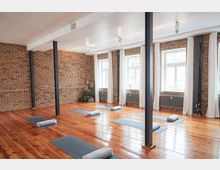 Ein heller Yogaraum mit Holzboden, freiliegenden Ziegelwänden und großen Fenstern mit weißen Vorhängen. Mehrere Yogamatten mit Kissen liegen gleichmäßig im Raum verteilt, während schwarze Stahlträger die Decke stützen.