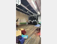 Spacious indoor studio or event room with wood-look flooring, an exposed brick wall, and a grid ceiling with fluorescent strip lights and hanging circular decorations. Green chairs line one wall, sheer fabric drapes hang on the walls, and boxes and equipment are scattered around the open floor.