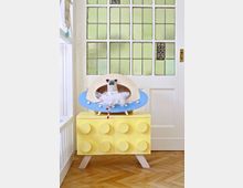 Eine helle Inneneinrichtung mit einer gelben Kommode, deren Schubladen wie große Legosteine gestaltet sind. Im Hintergrund ist eine weiße Tür mit matten Glasfeldern in grünlichen Tönen zu sehen, und auf der Kommode steht ein Modell eines UFOs mit einer kleinen Tierfigur.