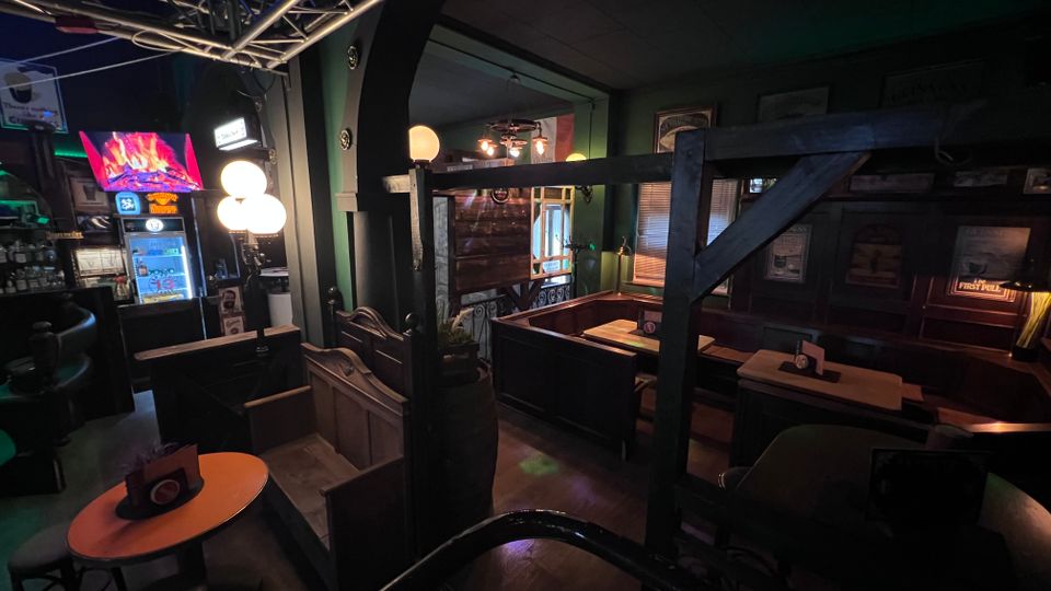 Innenansicht einer dunkel gestalteten Bar mit Holzvertäfelung, Nischen-Sitzbänken und kleinen Tischen entlang der Wände. Links stehen Spielautomaten und eine Lichtanlage mit Metallträgern, während warme Kugelleuchten und gerahmte Wandbilder den Raum beleuchten.