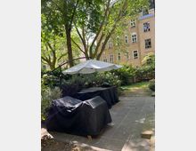 Gartenbereich mit gepflastertem Boden, einem großen weißen Sonnenschirm und abgedeckten Gartenmöbeln sowie einem Grill. Im Hintergrund sind Bäume und ein beige-farbenes Mehrfamilienhaus mit weißen Fenstern zu sehen.