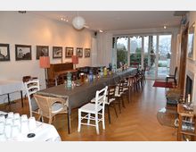 Großes Wohn- oder Veranstaltungszimmer mit langen Tischen, die mit Stühlen umgeben sind. An den Wänden hängen gerahmte Fotografien, und im Hintergrund sind große Fenster mit Blick auf einen Garten zu sehen.