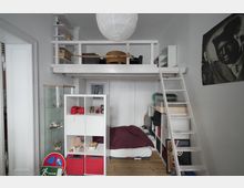 Kompakter Wohnraum mit Hochbett aus weißem Holz, einer Leiter und darunter einem Bett mit dunkelroter Bettdecke. Links stehen ein offenes Regal und eine Glasvitrine, rechts befinden sich weitere Regale mit Büchern und Gegenständen. An der Wand hängt ein großformatiges Schwarz-Weiß-Bild.