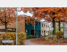 Reihenhäuser mit flachen Dächern in einem Wohngebiet, umgeben von herbstlich gefärbten Bäumen und Sträuchern. Ein Gehweg führt an den Gebäuden entlang, und ein Schild weist auf einen Privatweg hin.