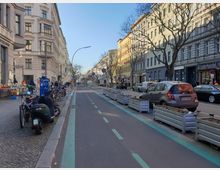Straßenszene in einer Stadt mit mehrstöckigen Wohngebäuden und Geschäften, darunter ein Lederwarenladen. Im Vordergrund befinden sich ein separater Fahrradweg mit Begrenzungen und geparkte Fahrräder sowie Mopeds, daneben Autos auf der Straße.
