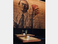 Holzvertäfeltes Café mit dunklen Sitzbänken und kleinen Holztischen, dekoriert mit bernsteinfarbenen Vasen, Zweigen und Teelichtern. Im Hintergrund ist eine große Wandillustration in Sepiatönen mit einem Mann und einer Frau zu sehen.