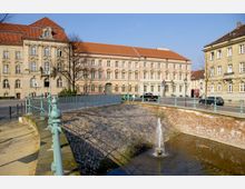 Historische Gebäude mit roten Ziegeldächern und verzierten Fassaden stehen an einer gepflasterten Straße mit parkenden Autos. Im Vordergrund befindet sich eine Wasseranlage mit einem kleinen Brunnen und einem Geländer aus Metall in mintgrüner Farbe.